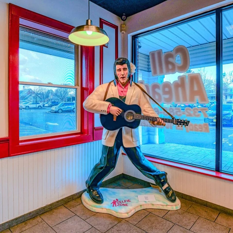Life-size Elvis Presley statue playing guitar inside The Burger Shop, with red window frames and blue sky.