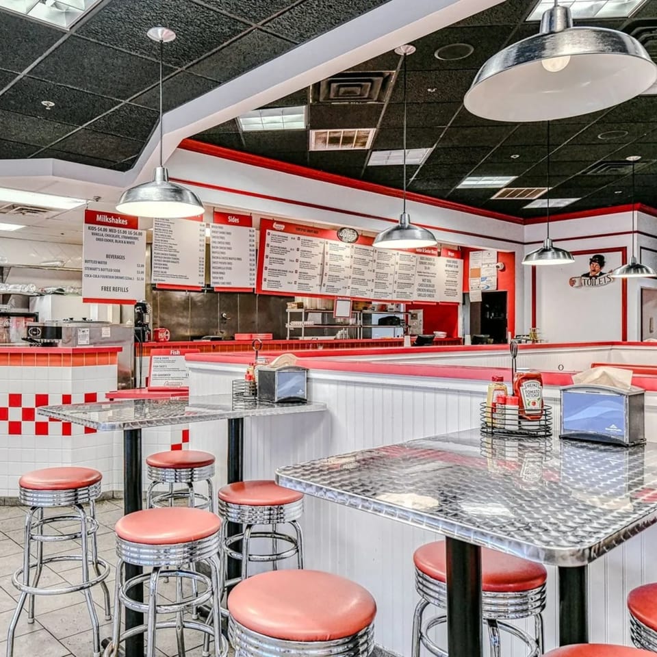 Interior of The Burger Shop, a fast-food restaurant with red and white checkered walls, chrome tables, and red stools.