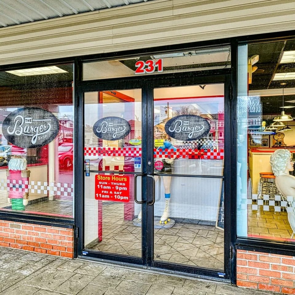 Exterior of The Burger Shop with store hours, checkered window decals, and a mannequin inside.