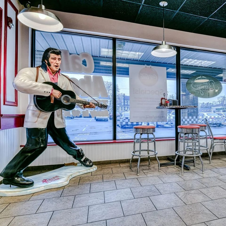 An Elvis Presley statue with a guitar stands in a retro diner with red stools and large windows.