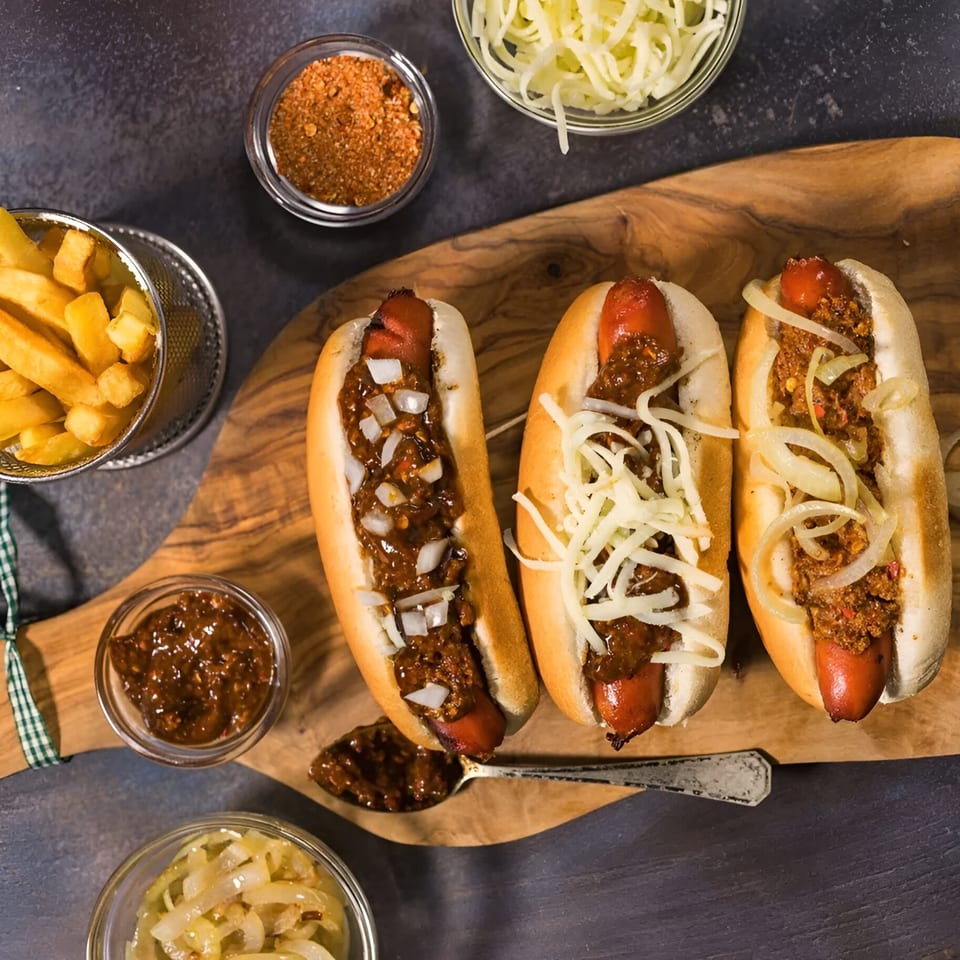 Three chili dogs with various toppings, fries, and condiments on a wooden board.